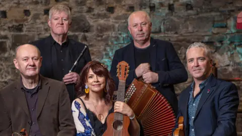 Tim Jarvis/Dervish A group of older looking musicians.
Left to right: a bald man in a brown jacket sitting, a grey haired man in a black jacket holding a black whistle, a woman with reddish brown hair, blue and yellow earrings and a blue shawl over a black vest top holding a brown ukulele, a bald man in a black jacket standing holding an accordion and a man sitting with grey short hair in a navy jacket with a brown fiddle under his arm.