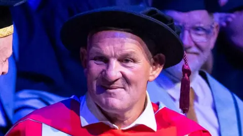 University of Hull/Mike Park Dean Windass is wearing a red and green gown and a black hat with a red thick tassel dangling on the right. He is in an arena and is smiling to the audience. There is an academic smiling behind him.