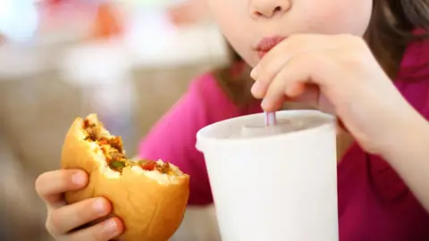 Getty Images A child in a pink T-shirt sipping a drink through a straw with a half-eaten burger in their other hand