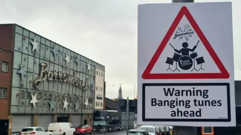 @ASignOfHumour A red triangle warning sign that reads "Warning banging tunes ahead" with a cartoon man playing a drum set. The sign is in front of Glasgow's Barowland sign.