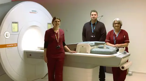 North Bristol NHS Trust Three members of staff, two in medical clothes, standing beside the donated MRI scanner. It has a tunnel to the left, with a bed to the right of it.