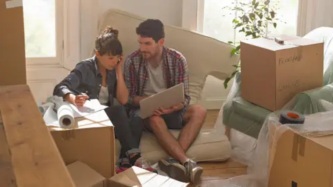 Getty Images A man and a woman sit on a cushion looking at a laptop computer surrounded by unpacked boxes.