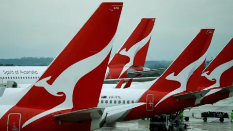 Reuters Four planes are lined up in a row on the tarmac of an aiport, each with the Qantas logo of a stylized white kangaroo on a red background emblazoned on the tail. 