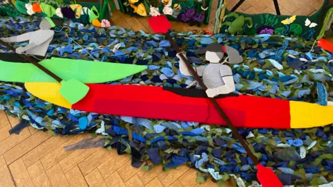 The Secret Society of Hertford Crafters Two kayker's made out of fabric, holding oars, on a river bed, also made out of fabric. The items are placed on a wooden floor.