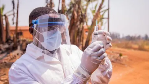 Getty Images A medic in full protective face mask and white clothing, standing outside, holding up a syringe with gloved hands