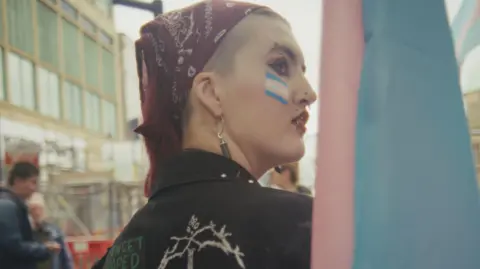 eden film Mason is at a rally and looking over his shoulder. He is wearing make-up and a dark-red bandana. He has a blue, pink and white transgender Pride flag painted on his face.