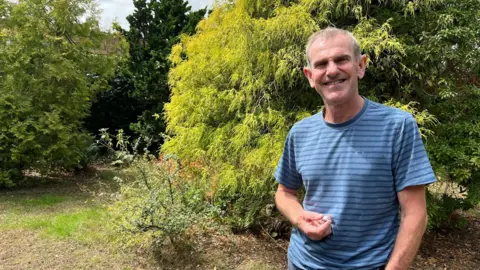 BBC Gregory Quinn stands in his front garden with a thick green hedge behind him. He is wearing a blue striped t shirt and is smiling at the camera. 
