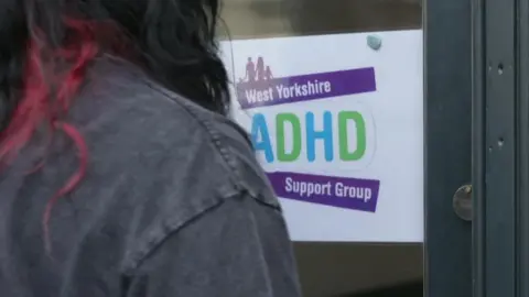 Beth Parsons/BBC A person with red and black hair and a black jacket, with their back to the camera, faces a white sign attached to a door. The sign says, in colourful writing, West Yorkshire ADHD Support Group.