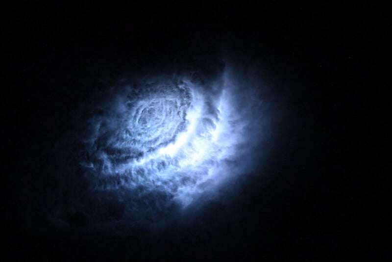 Bright, swirling blue and white light forms a spiral pattern against a pitch-black background, resembling a glowing galaxy or nebula in space.