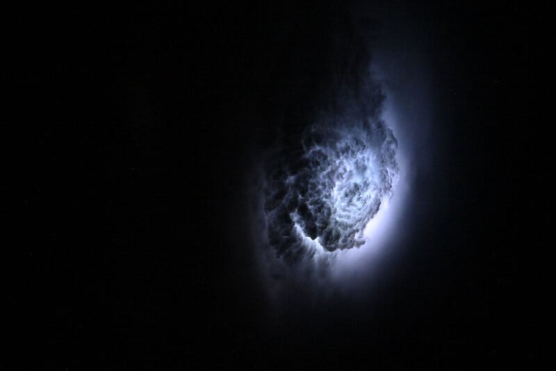 A glowing, cloud-like nebula with bright white and blue light at its center is surrounded by darkness, giving the appearance of a cosmic storm or a luminous cloud in deep space.