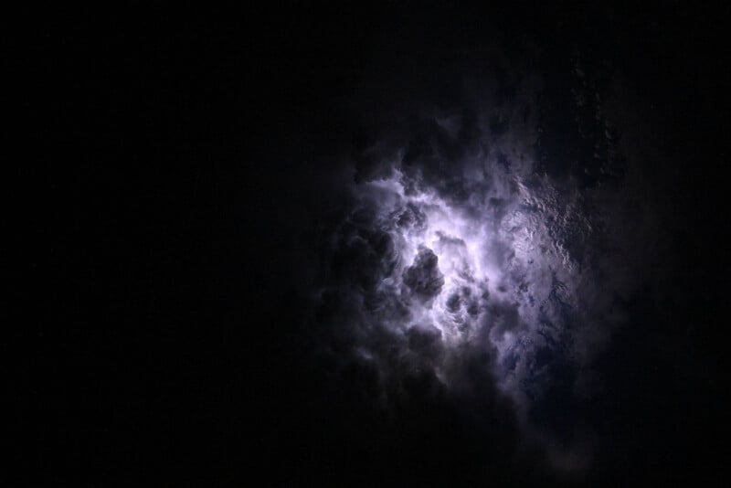Dark storm clouds illuminated from within by a bright flash of lightning, surrounded by deep shadows, creating a dramatic and intense atmosphere in the night sky.