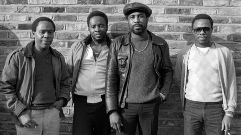 David Corio/Getty Images Black and white photo of B.B. Seaton of The Gaylads, Jackie Robinson of The Pioneers, A J Franklin of Chosen Few and Junior English standing in front of a brick wall