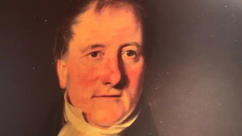 BBC A painting of Henry Duncan against a dark background. He has short brown hair and is wearing a white shirt and black robe. 