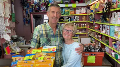 BBC Terry has short hair and is wearing a green checked shirt. He is standing with his arm around Orla who has short blonde hair and is wearing glasses and a blue and white striped top. They are both stood at the counter in the pet shop smiling at the camera.