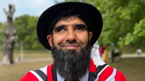 Elliot Deady/BBC A smiling Wali Noori looking straight at the camera. He is wearing graduation robes over a suit. He is standing on a grassy hill with threes in the background.