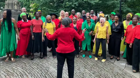 A group of people wearing red, green and yellow singing in a choir outside the church where Lawrence's funeral was being helpd