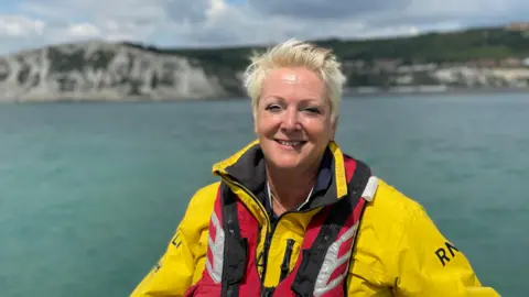 Simon Jones/BBC Paula Lain - a woman with short blonde hair in a yellow RNLI wetsuit and a red life jacket. The picture has been taken from a boat in the sea. In the background is the White Cliffs of Dover.