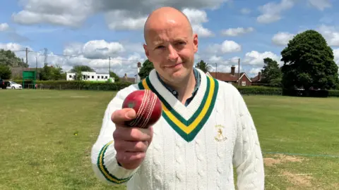 Matt Knight/BBC A head and shoulders picture of Rory Carlton, club secretary of Danbury Cricket Club, wearing his white cricket jumper and holding a red cricket ball on a cricket pitch.