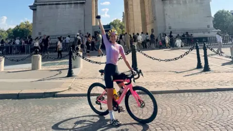 Amy Cycling Adventures Amy Hudson at the Arc de Triomphe in Paris. She is dressed in a pink and white cycling top, black shorts and a yellow helmet. She is sitting on her pink bike with her arm pointing in the air, with the base of the Arc de Triomphe behind her in light coloured stone.