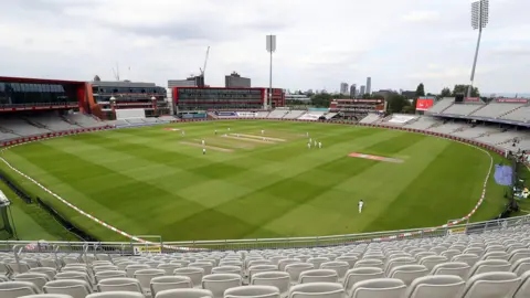 PA Media General view of the action on the pitch at Emirates Old Trafford ground in front of empty stands with floodlights the city skyline in the background.