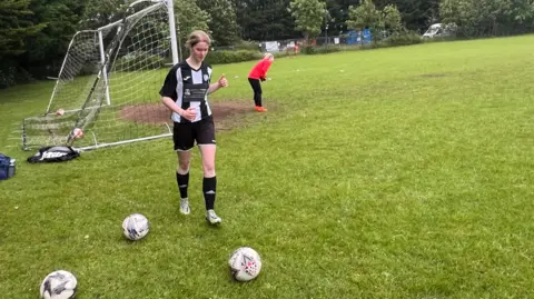 BBC A football player from Odd Down Women's AFC wearing a black and white striped top and black shorts goes towards a football near the goal on a pitch.