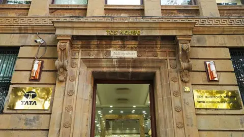 Getty Images The image is of the Mumbai headquarters of TCS. It is an ornate colonial building in brown stone, with golden plates of TATA and Tata Consultancy Services displayed on either side of the main door.