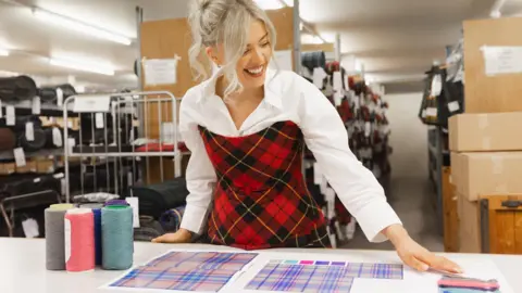 Glasgow 2026 Designer Siobhan Mackenzie, a young blonde haired woman, leans over her white workbench where her tartan design is laid out, with samples of the thread colours. She wears a white shirt and a stylish tartan dress.