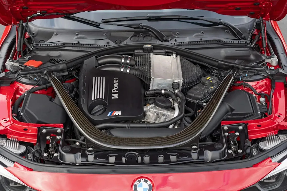 engine bay view showing inline six engine with carbon strut brace and black m power cover under raised hood.