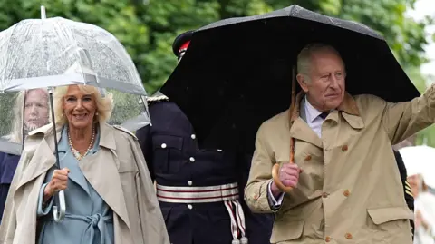 PA Media Queen Camilla in a blue dress with a beige trench coat holding an umbrella walking towards the camera, with King Charles III in a brown coat holding a black umbrella and waving.