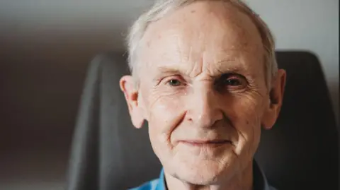 Association of Optometrists Headshot of an elderly man, with white hair and wearing a blue shirt and standing in front of a fireplace in a sitting room