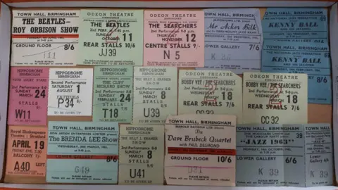 Richard Winterton 17 pastel-coloured ticket stubs laid out in a cardboard box for bands including The Beatles, The Searchers and Cliff Richard, all priced in shillings and pence.