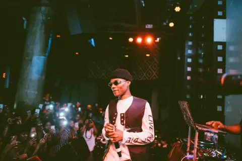 Oliver Akinfeleye Wizkid in round sunglass and a brown beanie, cream sweatshirt and brown waistcoat, holds a microphone on a small stage. To his right can be seen a small mixing desk and to left a group of mainly female fans taking photos of him with their phones.