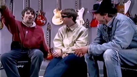 'The Beat' Fuji Television, 1994 Liam and Noel Gallagher sit with Gary Crowley in a 1994 TV interview, surrounded by guitars hanging on the wall behind them. Liam gestures animatedly, Noel wears sunglasses, and Crowley laughs in a denim jacket and cap.