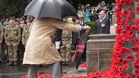 PA Media King Charles wearing a brown coat and holding a black umbrella lays at wreath a war memorial decorated with red poppies. Soldiers and the public line the background of the photo.