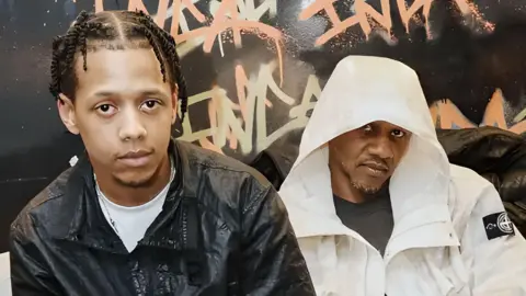 @vannibassett ML and Giggs sit on a sofa, ML leaning forward in front of his dad. ML wears a dark short sleeved shirt while Giggs wears a long sleeved white jacket with the hood up. They're pictured in front of a dark wall that's been graffitied. 