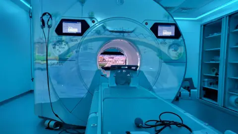 Alder Hey Children's Hospital The scanner has two screens on the outside and a screen can also be seen inside at the back. A table has a support for the patient's head. There is relaxed blue lighting in the room.