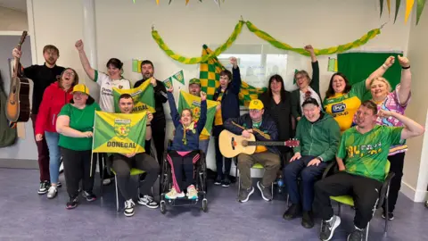 The group from the Community Inclusion Hub who star in the Big Jim's Gonna Do It Again music video. They are sitting in a row and many of them are wearing Donegal county colours or holding Donegal flags in yellow and green. Some are holding guitars and some are cheering.
