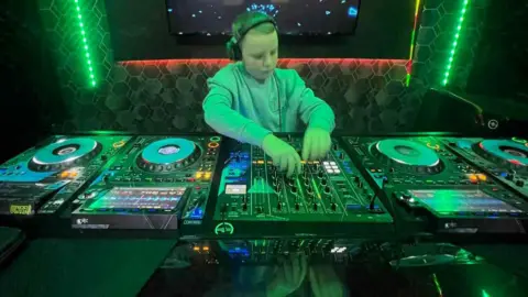 Kerry O'Kane Fionn is seen in a grey jumper with a pair of headphones on. 
He is at a DJ booth with a number of green lights behind him. 
He is mixin and staring at the decks. 