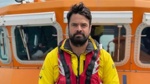 Simon Jones/BBC Dan Sinclair - a man with black hair, a black beard and a black moustache. He is wearing a yellow RNLI wetsuit and a red life jacket. The picture has been taken from a boat in the sea.