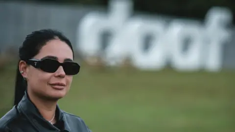 BBC Hannah Laing, in dark sunglasses, her dark hair in a ponytail and wearing a black leather jacket, stands with a large "Doof" sign behind her in Camperdown Park