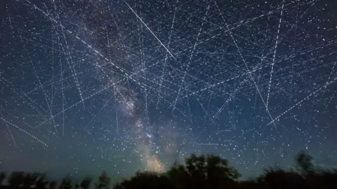 Getty Images An image of the night sky taken on a long exposure which shows it criss-crossed with dotted streaks from satellites moving through the sky. At the bottom of the image a dark line of trees is silhouetted while you can also see the stars and bright light of the milky way in the image.