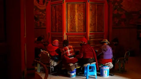 Xiqing Wang/ BBC Tibetan women sit by a prayer wheel inside a monastery
