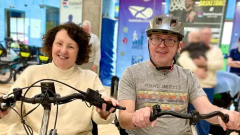 Easterhouse Phoenix Tony Deeney sits on a dual side by side tandem bike with his wife, Teresa. Tony holds onto the handlebars wearing a grey t-shirt with a grey helmet. He is smiling while looking at the camera. Teresa has short brown hair wears a cream jumper and holds onto the handlebars. She is smiling at the camera.