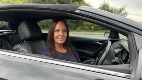 BBC Caroline O'Brien sitting in her car looking directly at the camera. She is wearing a blue top. She is visible from the shoulders up and is sitting in the driver's seat with the steering wheel in front of her. There is a road and foliage in the background.