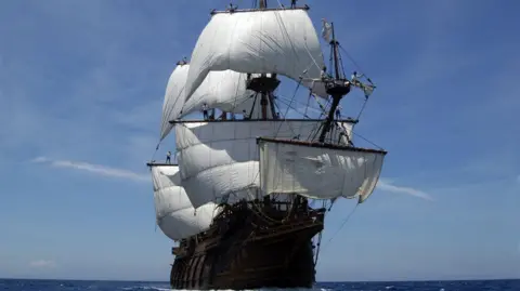 Bristol Harbour Festival A historic wooden ship with about half a dozen large white sails billowing in the wind. The ship is on the open ocean, with no land visible on the horizon. It is a sunny day, with a clear blue sky above it.