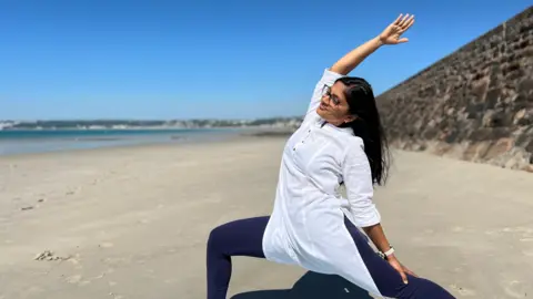 BBC Rashmi is holding a lunging pose on a beach as she wears a white light top with dark blue leggings