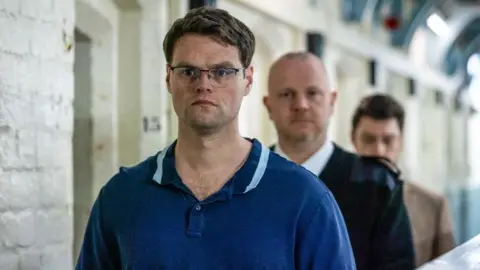 BBC/LA Productions/Kerry Spicer This candid still from the drama shows Joe, played by Bobby Schofield, walking along the hall in prison. There is a prison officer in uniform walking behind him. 