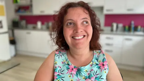 A person  sits in a kitchen with white cabinets and a pink wall. They are wearing a dress with a floral pattern of pink flowers and green leaves. Various kitchen items are visible on the counter.