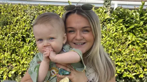 Handout A blonde woman with sunglasses on her head holding a blonde baby in a green shirt. Behind them is a green bush.