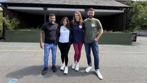 Sergio and Alex Aguiar and Lauren and Ben King are posing for a photograph in the middle of the playground at Churchtown Primary School. They are standing close together and have their arms around each other. 
